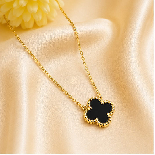 A gold chain necklace with a four-leaf clover pendant featuring a black center and a beaded gold border, displayed on a beige satin fabric background.