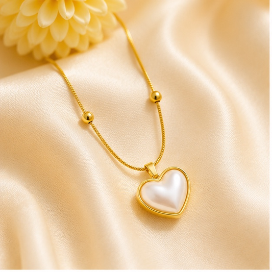 A gold heart pendant necklace with a white faux-pearl center, displayed on draped champagne satin fabric with a decorative flower in the corner.